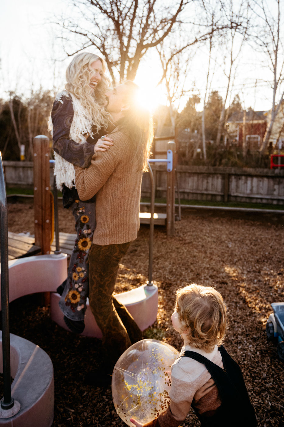 A joyful moment captured at a playground where a couple shares a loving embrace while sunlight filters through the trees, and a child holding a large clear ball looks on with curiosity.