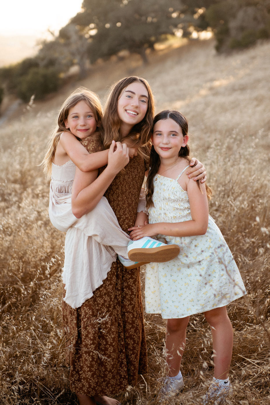 Three girls smiling in a dry, grassy field. One girl is being carried. They wear floral dresses in earthy tones, conveying a joyful mood.
