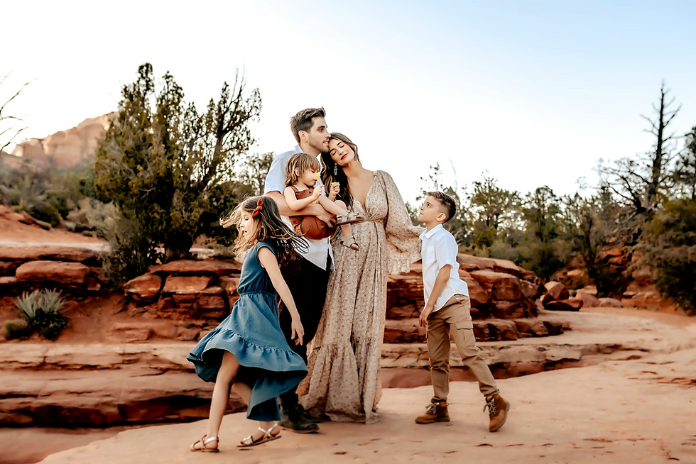 Family of five poses on rocky terrain, surrounded by trees. The mother holds a child. Warm colors and serene mood in the desert setting.