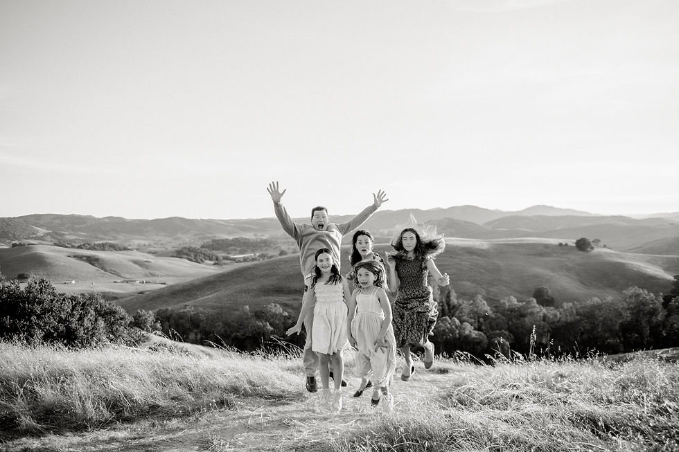 Family of six joyfully jumps in a grassy field with rolling hills in the background. Black and white photo with a cheerful, lively mood.