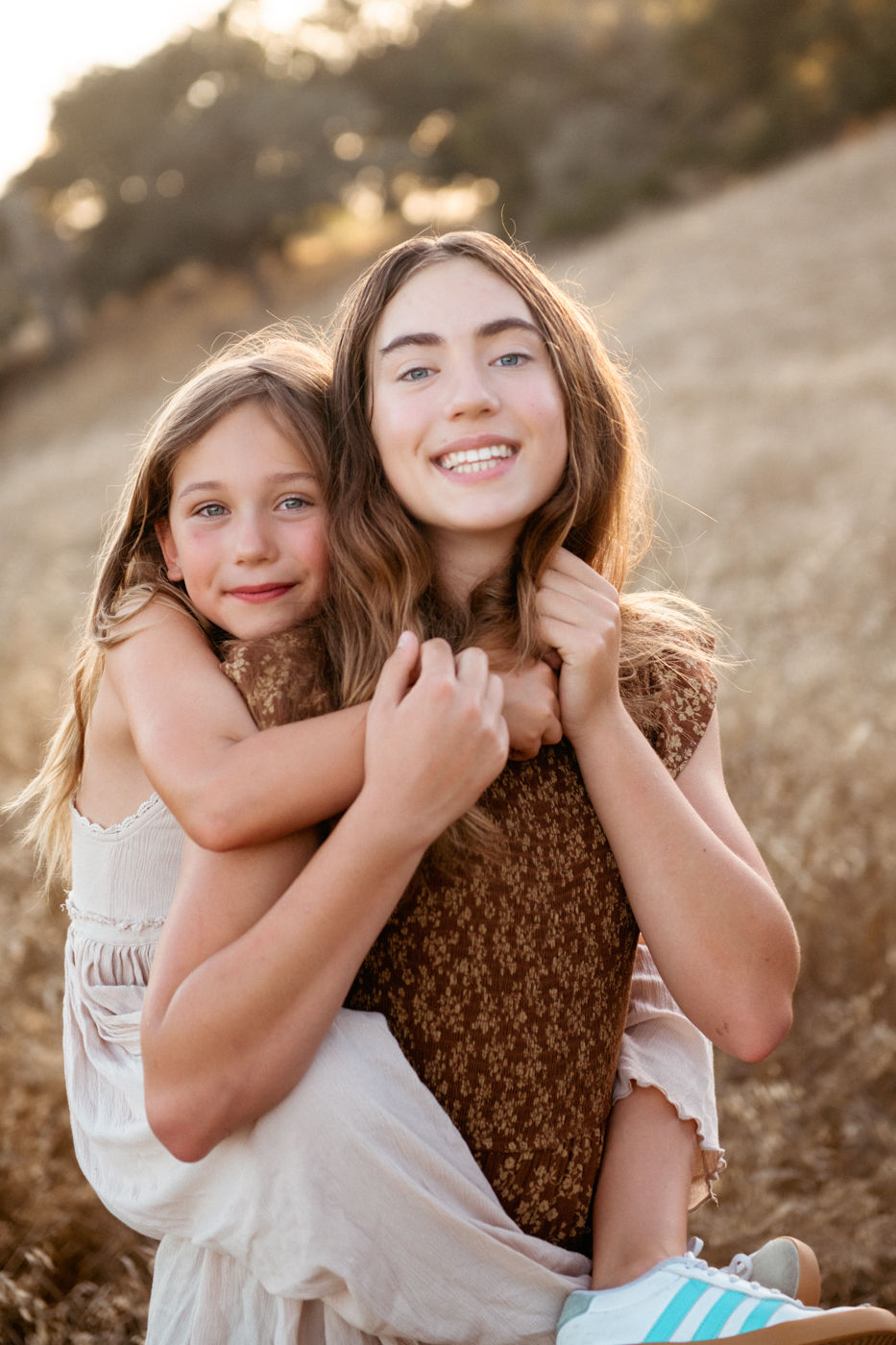 Two smiling girls in a field, one giving a piggyback ride. Brown patterned dress, cream outfit, striped sneakers. Warm, sunny background.