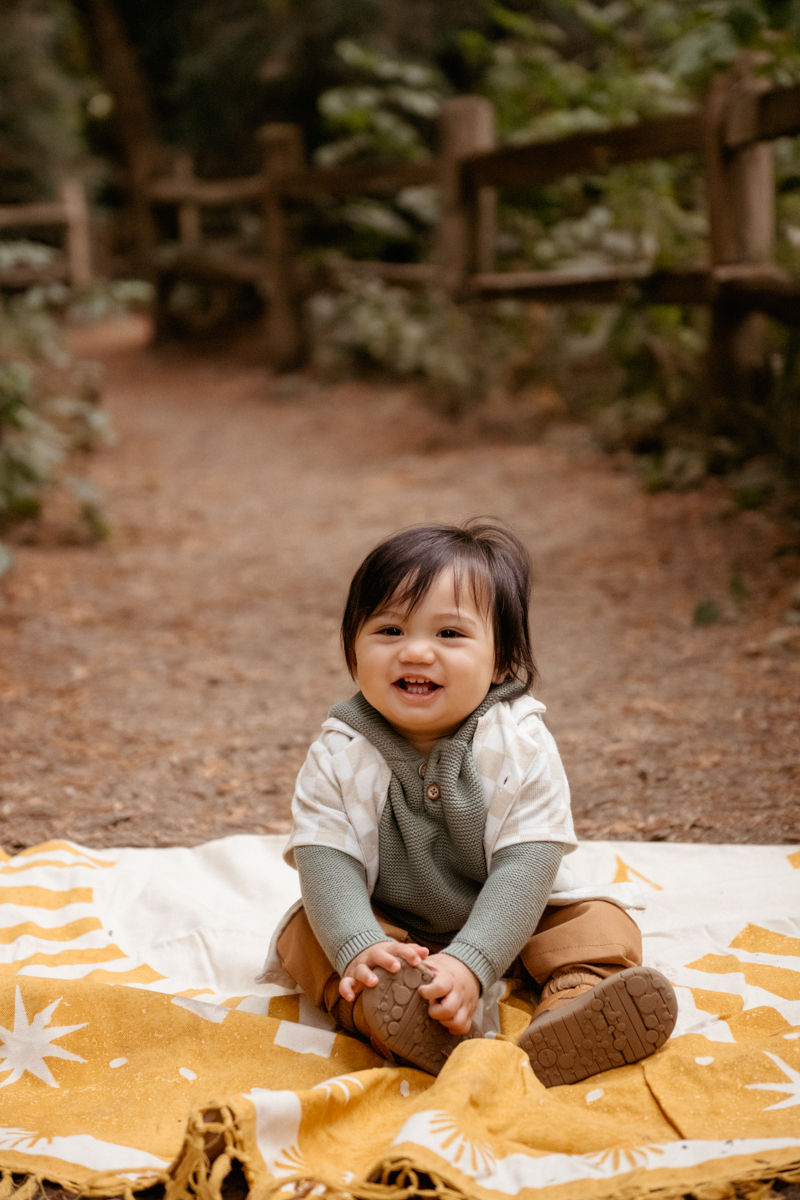 Smiling baby sits on a yellow-patterned blanket in a forest path, wearing a green sweater and brown pants. Warm, cheerful atmosphere.