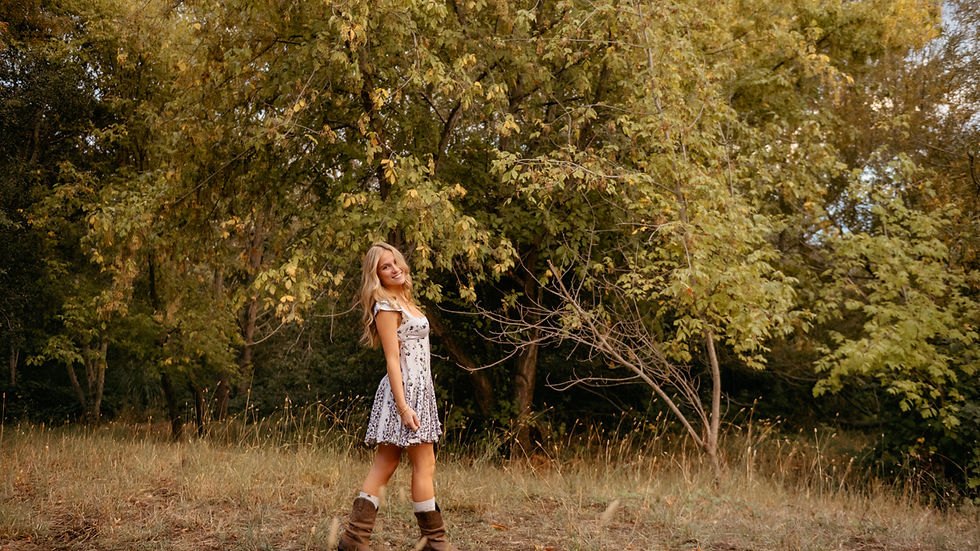 Woman in a floral dress and boots stands smiling in a sunlit forest clearing, surrounded by green foliage and tall grass.