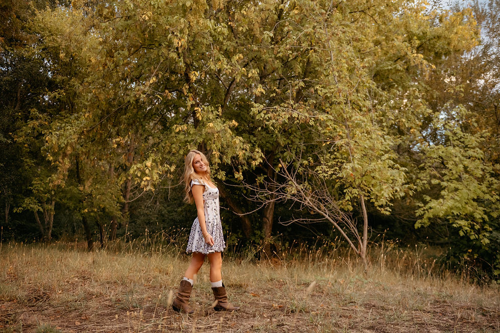 Woman in a floral dress and boots stands smiling in a sunlit forest clearing, surrounded by green foliage and tall grass.