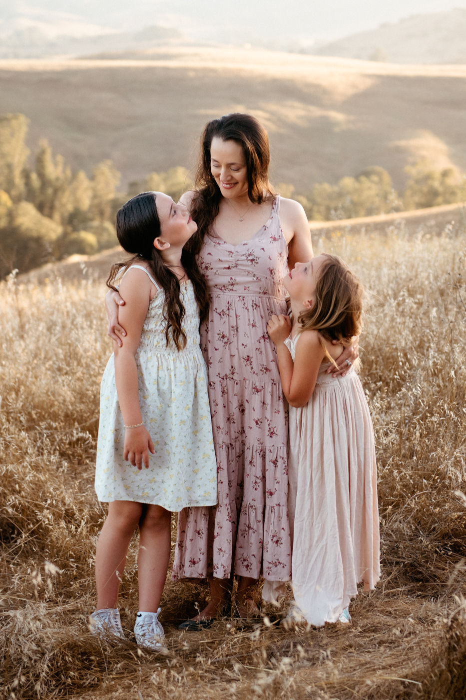 A woman and two girls in floral dresses stand in a sunlit field, smiling at each other. Hilly landscape in the background.
