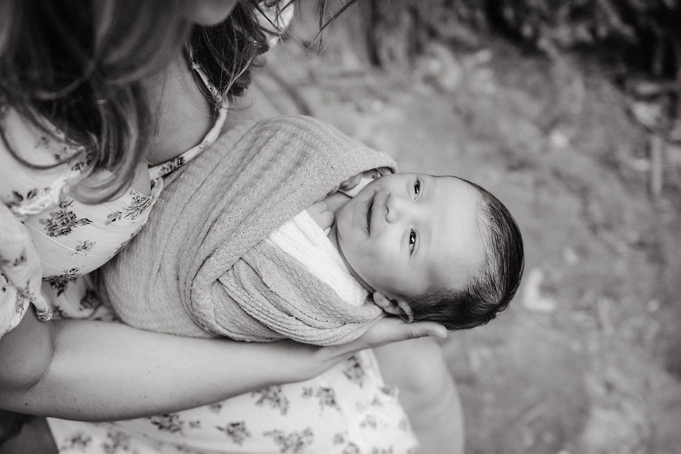 A woman in a floral dress cradles a smiling baby wrapped in a blanket. Black and white image, outdoors, evokes tenderness and warmth.
