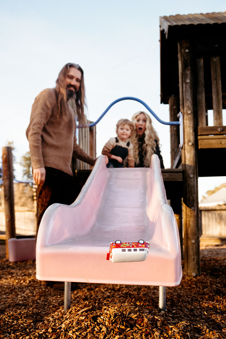 A family enjoys a playful moment at the playground, with a toy van racing down a pink slide, while they watch with excitement from above.