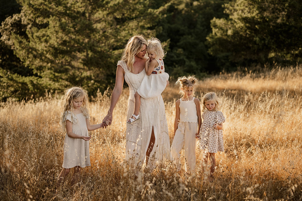 Woman with four young girls in a sunlit field, all wearing light dresses. Warm, serene atmosphere with trees in the background.