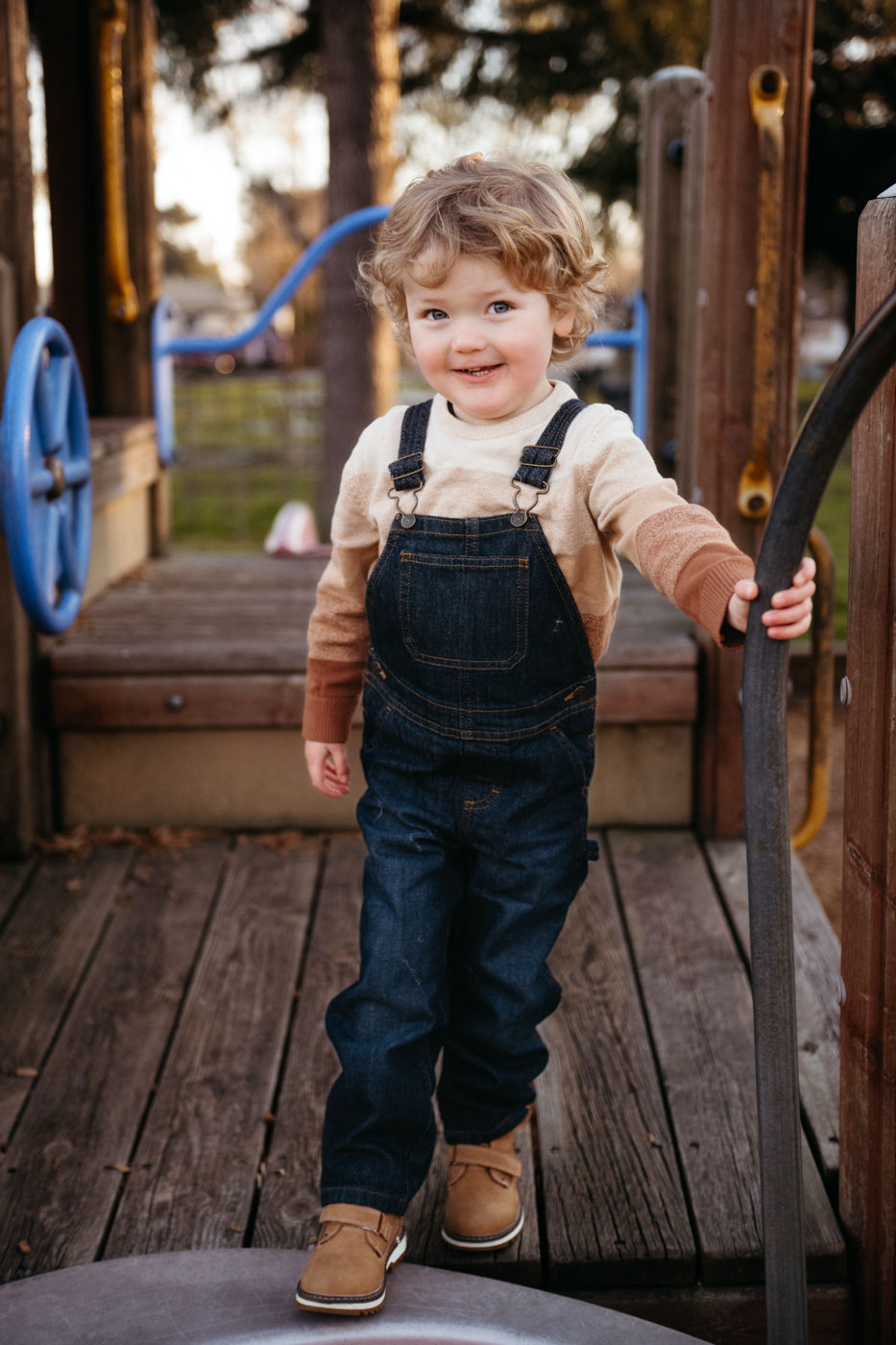 A joyful child in overalls beams with delight while exploring a wooden play structure on a sunny day at the park.