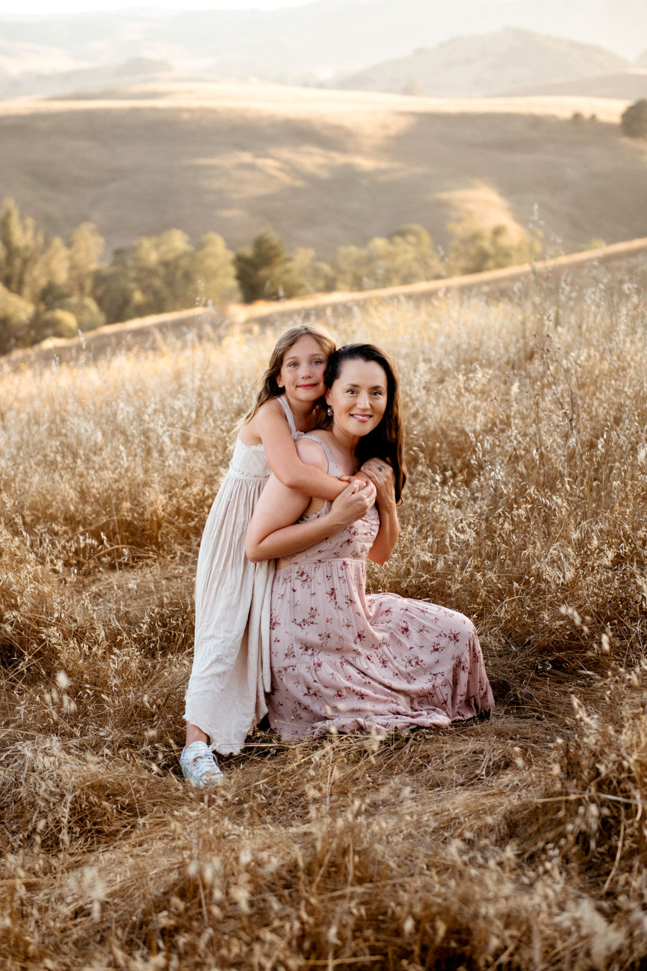 A woman and child in floral dresses embrace in a sunlit field, surrounded by golden grass and rolling hills. Warm, joyful mood.