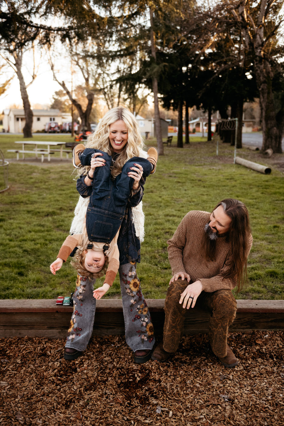 A joyful family moment unfolds in the park as a woman playfully lifts a child upside-down, while a man watches with a smile, surrounded by nature.