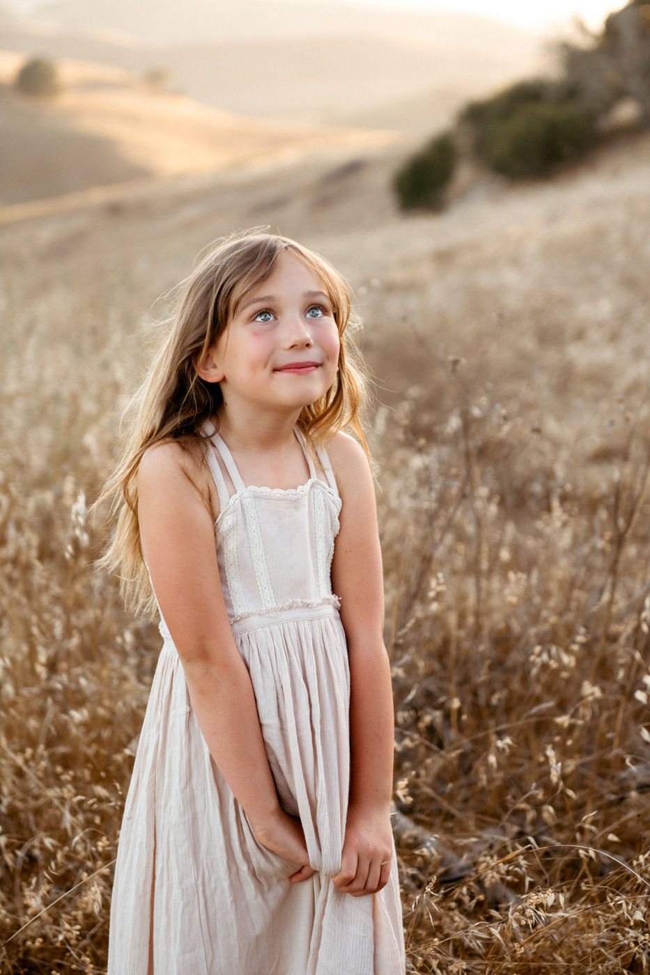 Young girl in a white dress smiles while standing in a golden field. Rolling hills and soft sunlight create a serene, dreamy atmosphere.
