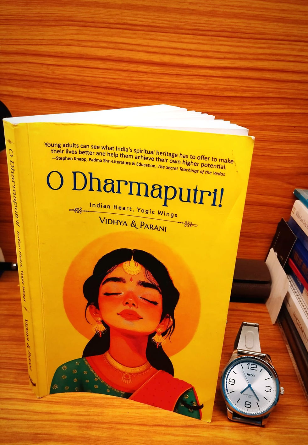 Yellow book cover, "O Dharmaputri!" with a serene illustration on a wooden surface, accompanied by a silver wristwatch. Books are in the background.