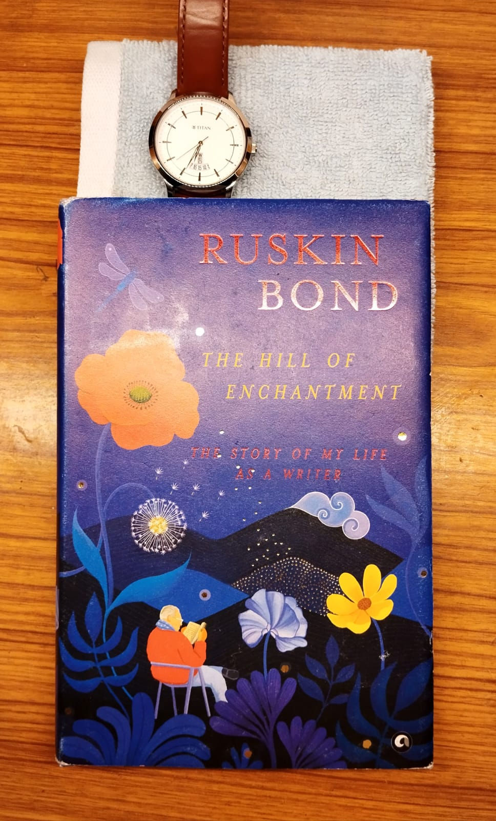 A book titled "The Hill of Enchantment" with a blue floral cover lies on a towel. A watch with a brown strap is placed above it.