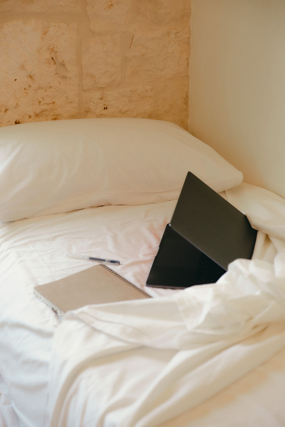 Laptop, Notizbuch und Stift auf einem hellen Bett als Beispiel für mobiles Arbeiten innerhalb der eigenen Wohnung.