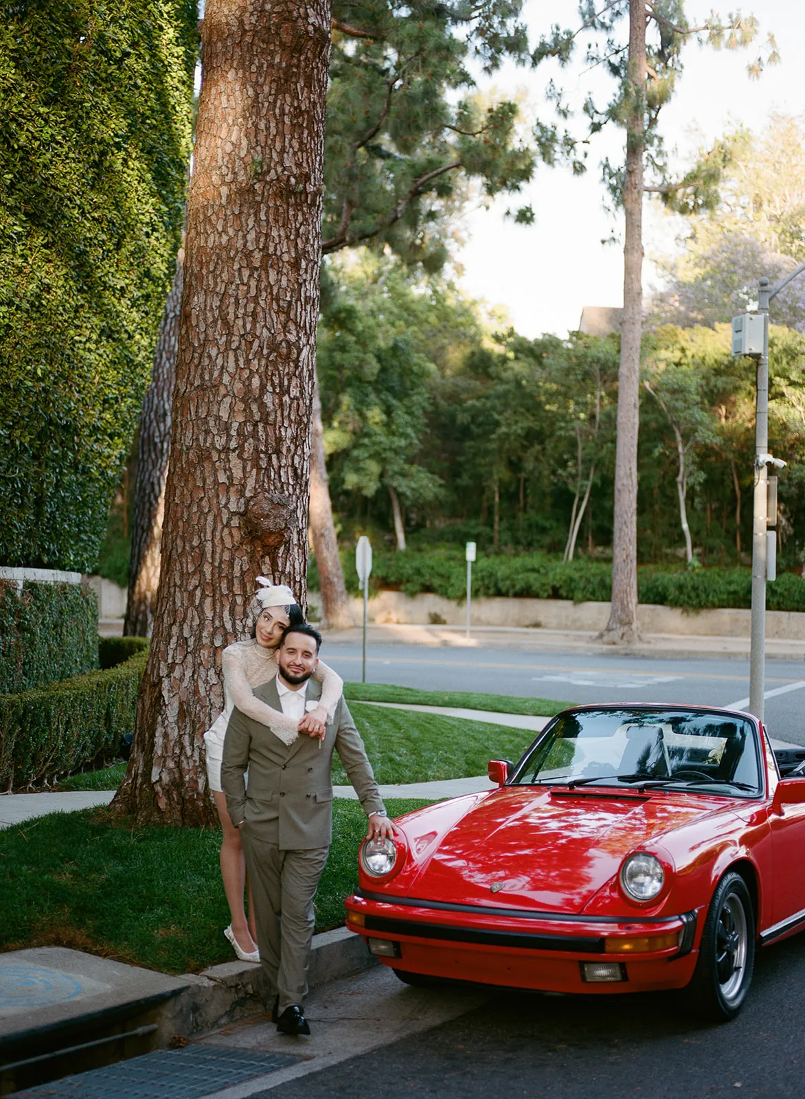 Couple embracing beside a large tree and vintage red Porsche 911 on a quiet Beverly Hills street, bride in short lace wedding dress and birdcage fascinator, groom in olive double-breasted suit, lush hedgerow behind them