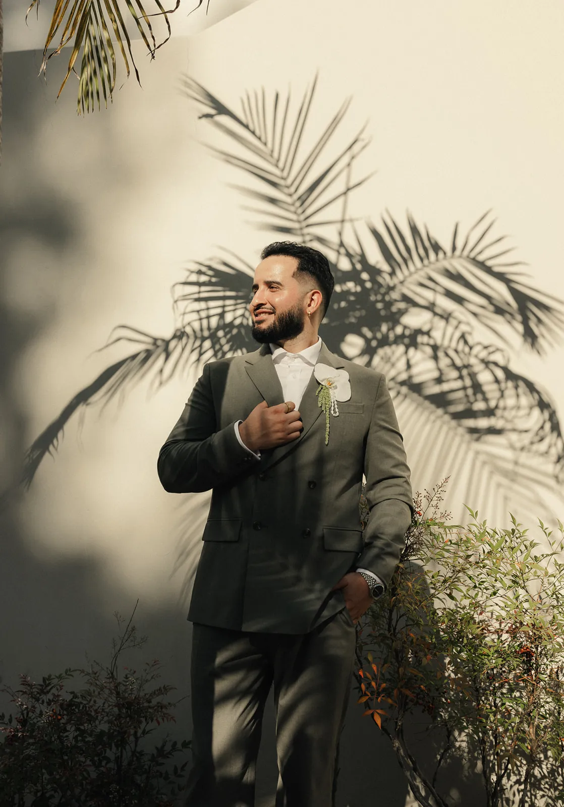 Groom portrait against white wall with dramatic palm tree shadow at Beverly Hills City Hall, olive double-breasted suit with white orchid boutonniere, smiling and looking away from camera, Beverly Hills courthouse wedding photography by Rene Porto