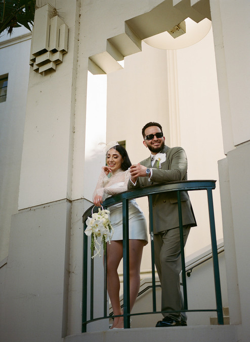 Couple on the curved balcony of Beverly Hills City Hall during a courthouse elopement, bride in short white lace dress with birdcage fascinator holding cascading white orchid bouquet, groom in olive suit with sunglasses, shot on film by Los Angeles wedding photographer Rene Porto