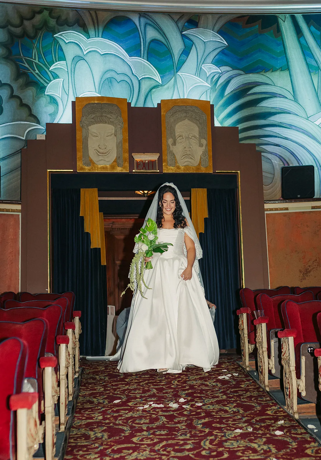 Bride walking down the aisle of the Avalon Theatre alone, veil and tropical bouquet, John Gabriel Beckman murals flanking the stage at the Catalina Island Casino wedding