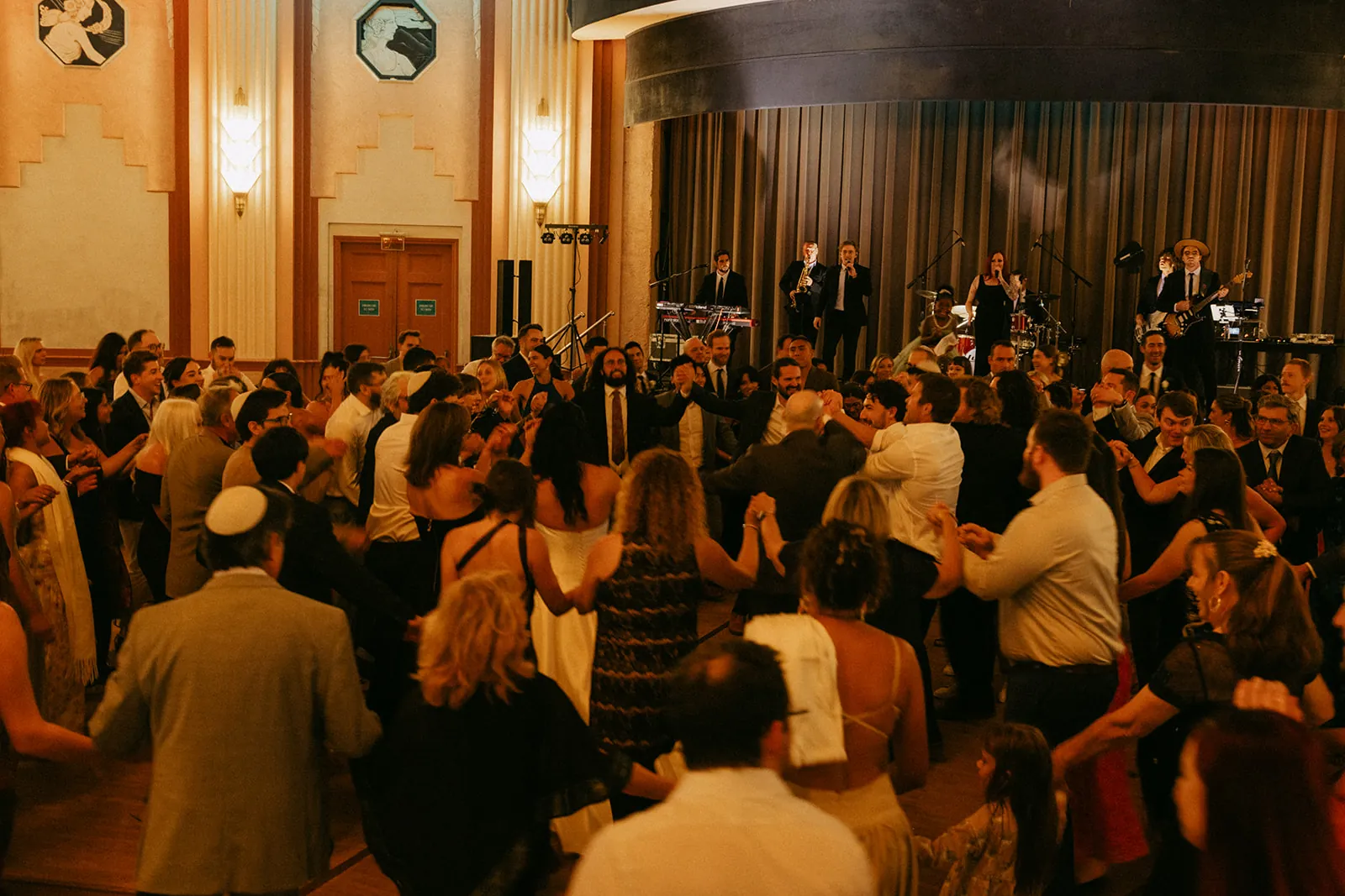 Full dance floor during the hora at a Catalina Island Casino Ballroom wedding reception, live band performing on stage and guests filling the room
