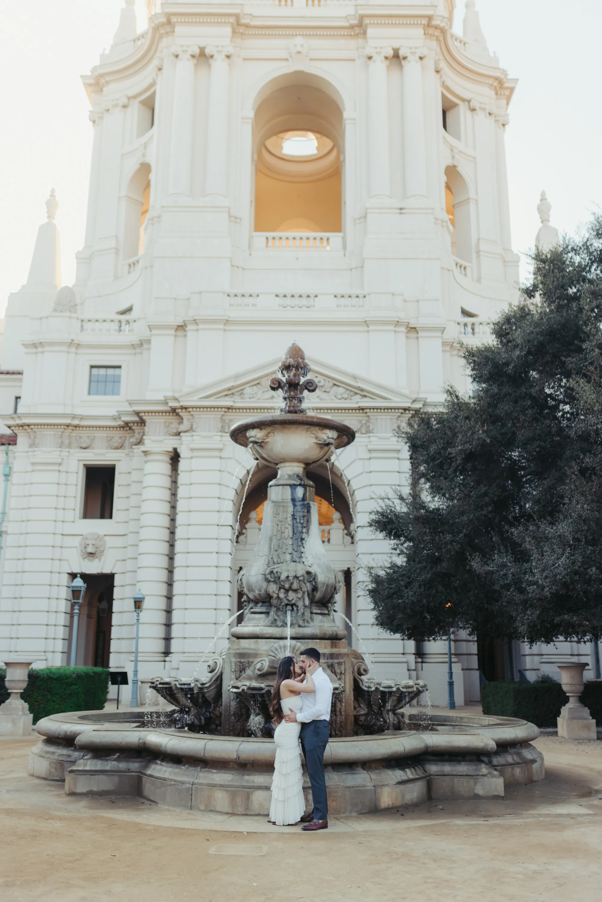 engagement photo outfits at Pasadena City Hall, couple in white fitted dress and navy trousers embracing in front of the ornate fountain with the City Hall dome in the background