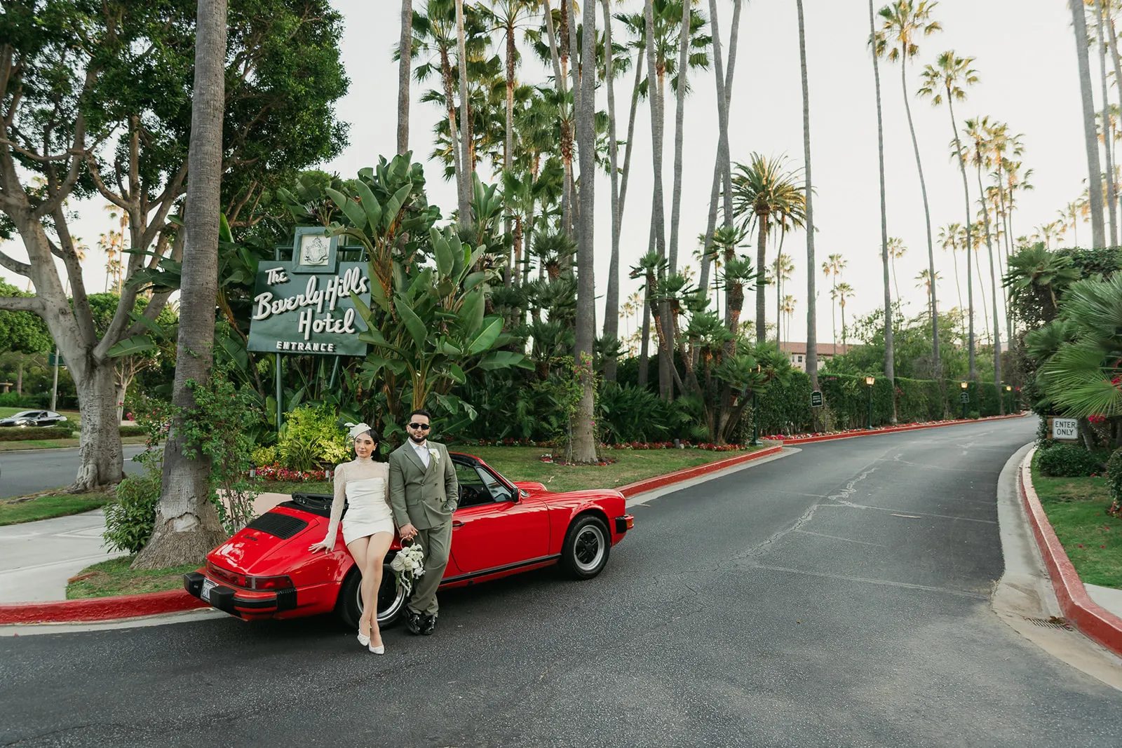 Wide portrait of couple leaning against a vintage red Porsche convertible in front of the Beverly Hills Hotel entrance sign, palm-lined driveway behind them, bride in short lace wedding dress and fascinator, groom in olive suit and sunglasses