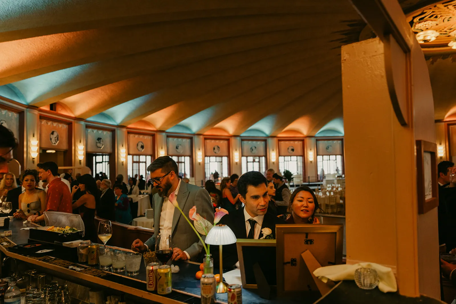 Guests at the Casino Ballroom bar through the pass-through window at a Catalina Island Casino wedding reception, amber and teal uplighting throughout the art deco room