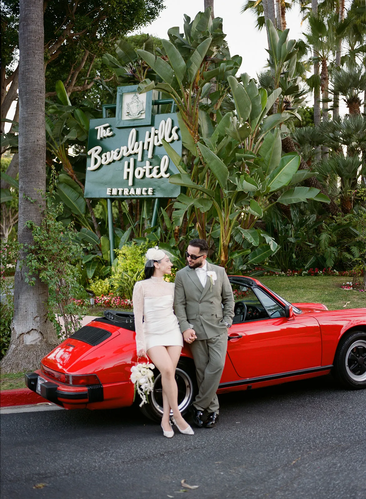 Couple in front of the Beverly Hills Hotel entrance sign leaning on a red Porsche after their courthouse elopement