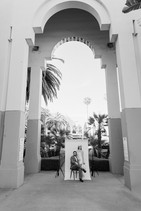 Wide black and white shot of couple posed against a white studio backdrop set up between the architectural arches of Beverly Hills City Hall, groom seated on stool with bride standing beside him, palm trees and Beverly Hills skyline visible through the arch behind them