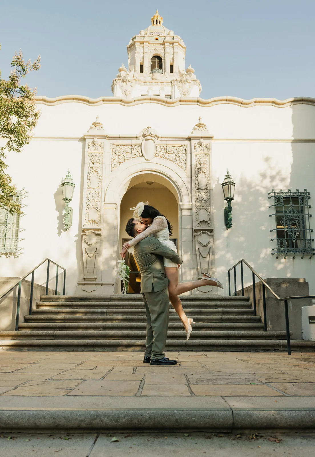 Groom lifting bride in front of the Beverly Hills City Hall entrance with gold dome tower above