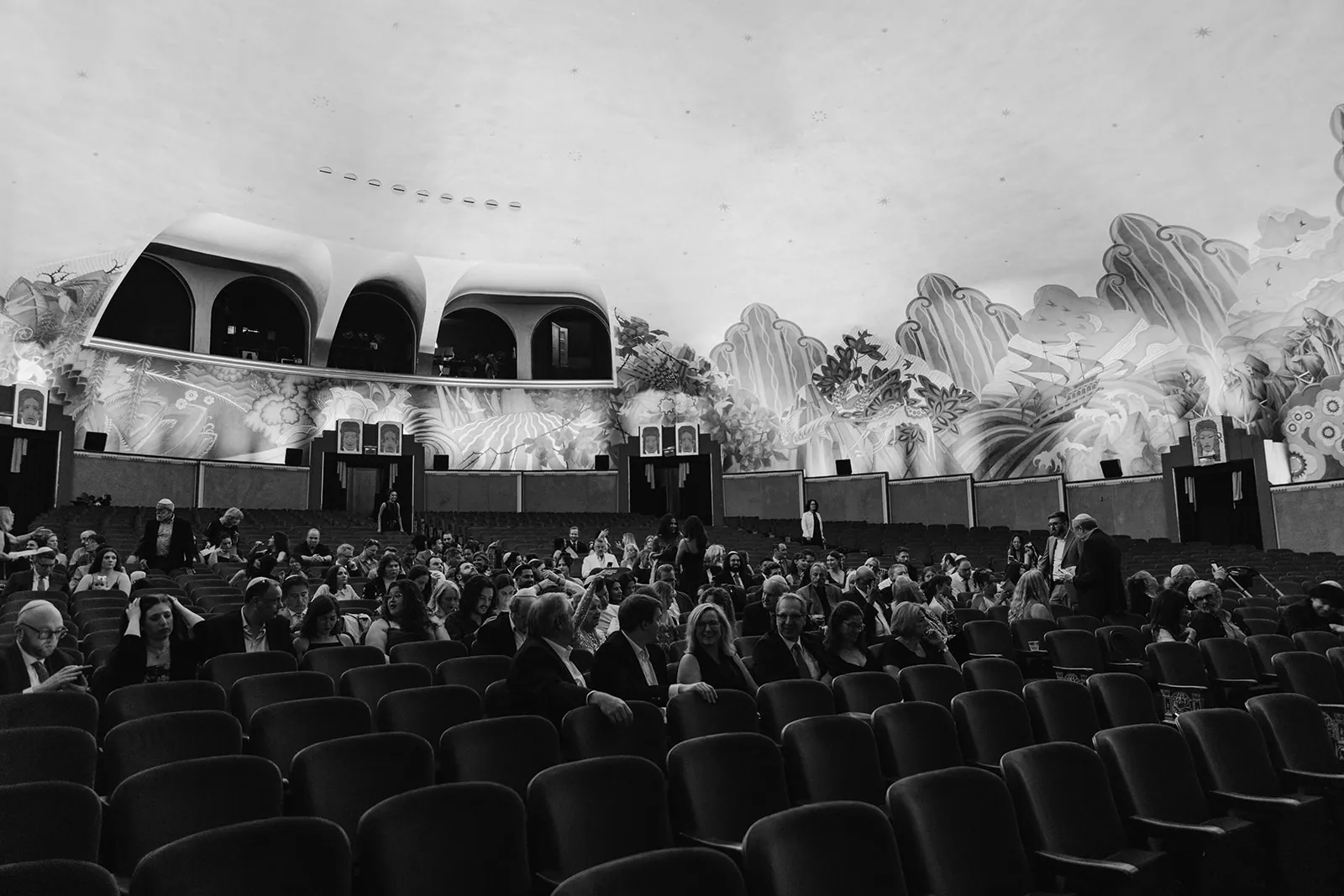 Wide shot from the stage of the full crowd seated in the Avalon Theatre at the Catalina Island Casino for the wedding ceremony, John Gabriel Beckman murals visible along the walls, black and white