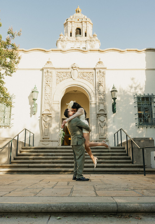 Groom lifting bride in a kiss in front of the ornate Beverly Hills City Hall facade and tower during their courthouse wedding, bride in short white lace dress and fascinator, groom in olive double-breasted suit, photographed by Los Angeles wedding photographer Rene Porto