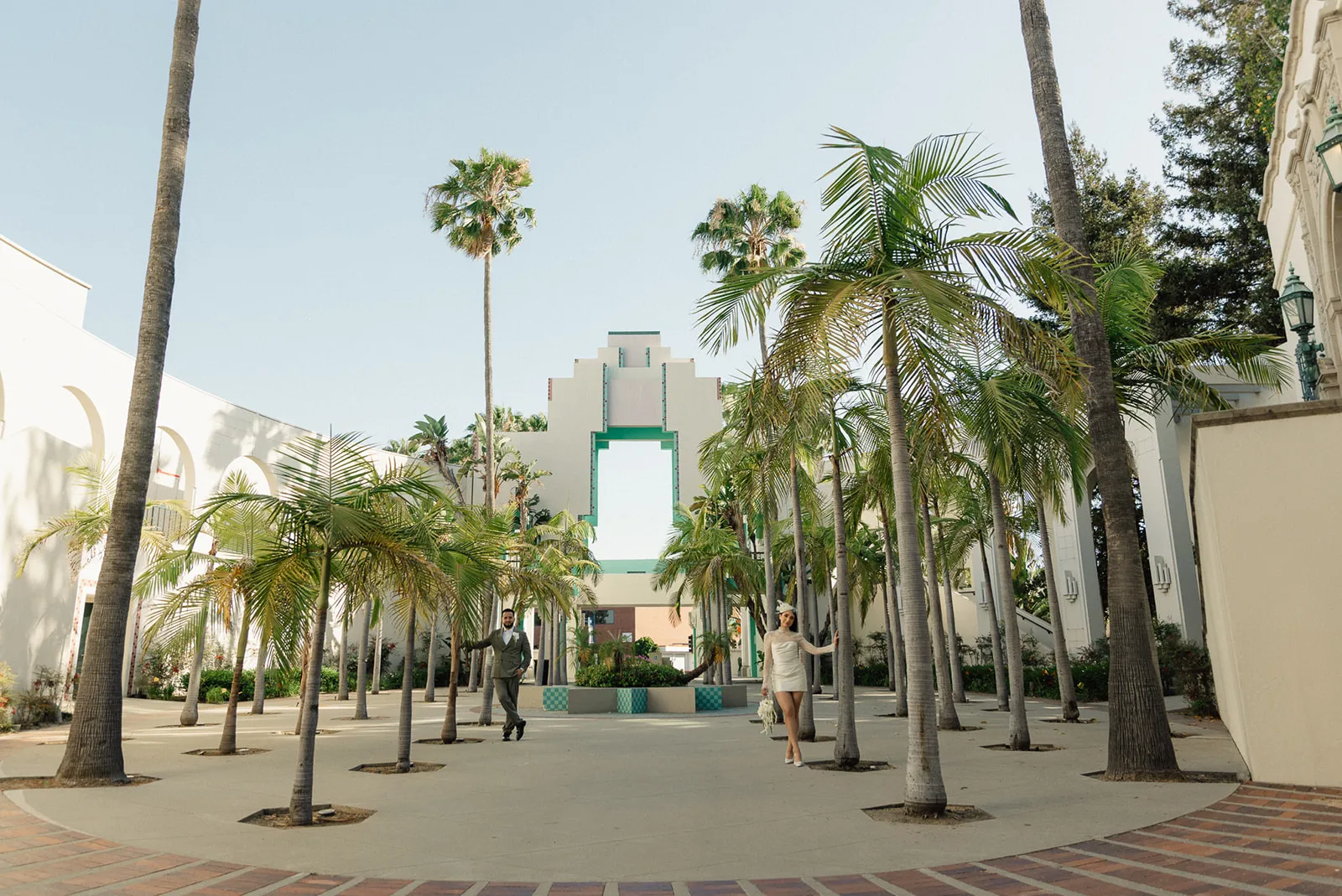 Wide shot of couple walking through the palm-lined civic center plaza at Beverly Hills City Hall, Art Deco tower visible in the background, bride in short white lace dress and fascinator, groom in olive suit, Beverly Hills courthouse wedding photography by Rene Porto