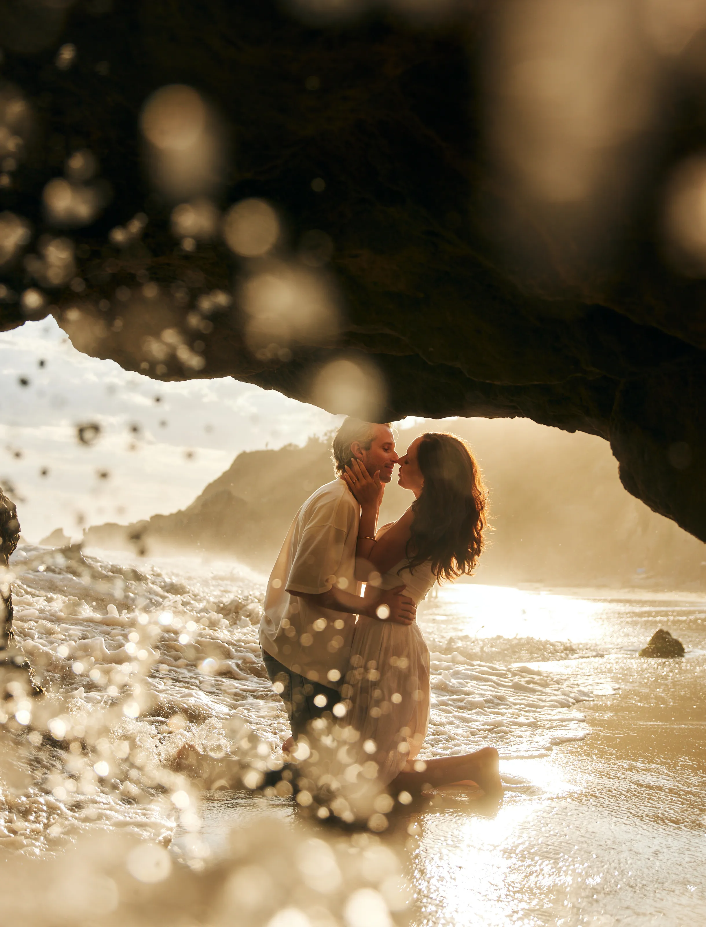 engagement photo outfits at El Matador Beach Malibu, couple in cream and white outfits kissing inside a sea cave with golden ocean backlight