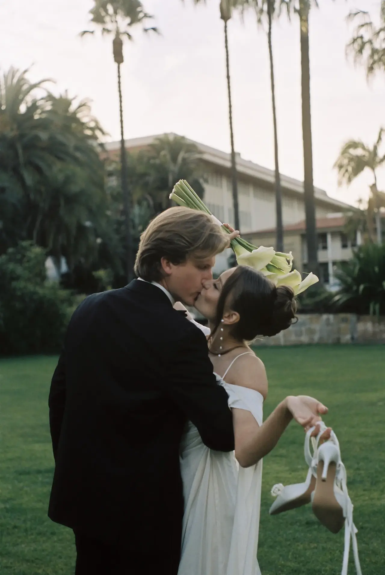 A bride in a white slip dress kisses her groom on the lawn as she raises a calla lily bouquet above them and holds her heels in her other hand, tall Canary Island palms and a California coastal venue behind them, shot on 35mm film by Los Angeles film wedding photographer Rene Porto