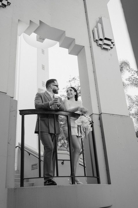 Black and white photo of couple on the curved balcony of Beverly Hills City Hall framed by Art Deco columns, bride in short lace wedding dress holding cascading white bouquet, groom in double-breasted suit and sunglasses, Beverly Hills elopement photography by Rene Porto