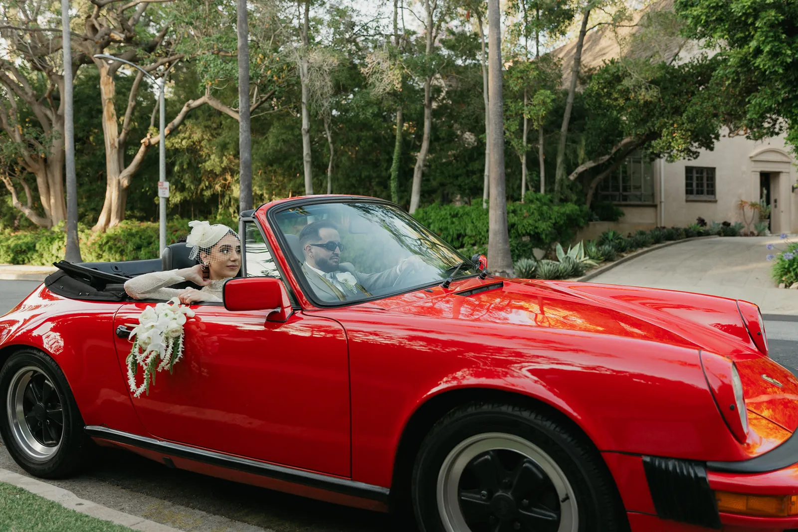 Couple driving through Beverly Hills streets in a vintage red Porsche convertible after their courthouse elopement, bride in lace dress and birdcage fascinator with white orchid bouquet draped over the door, groom in olive suit and sunglasses