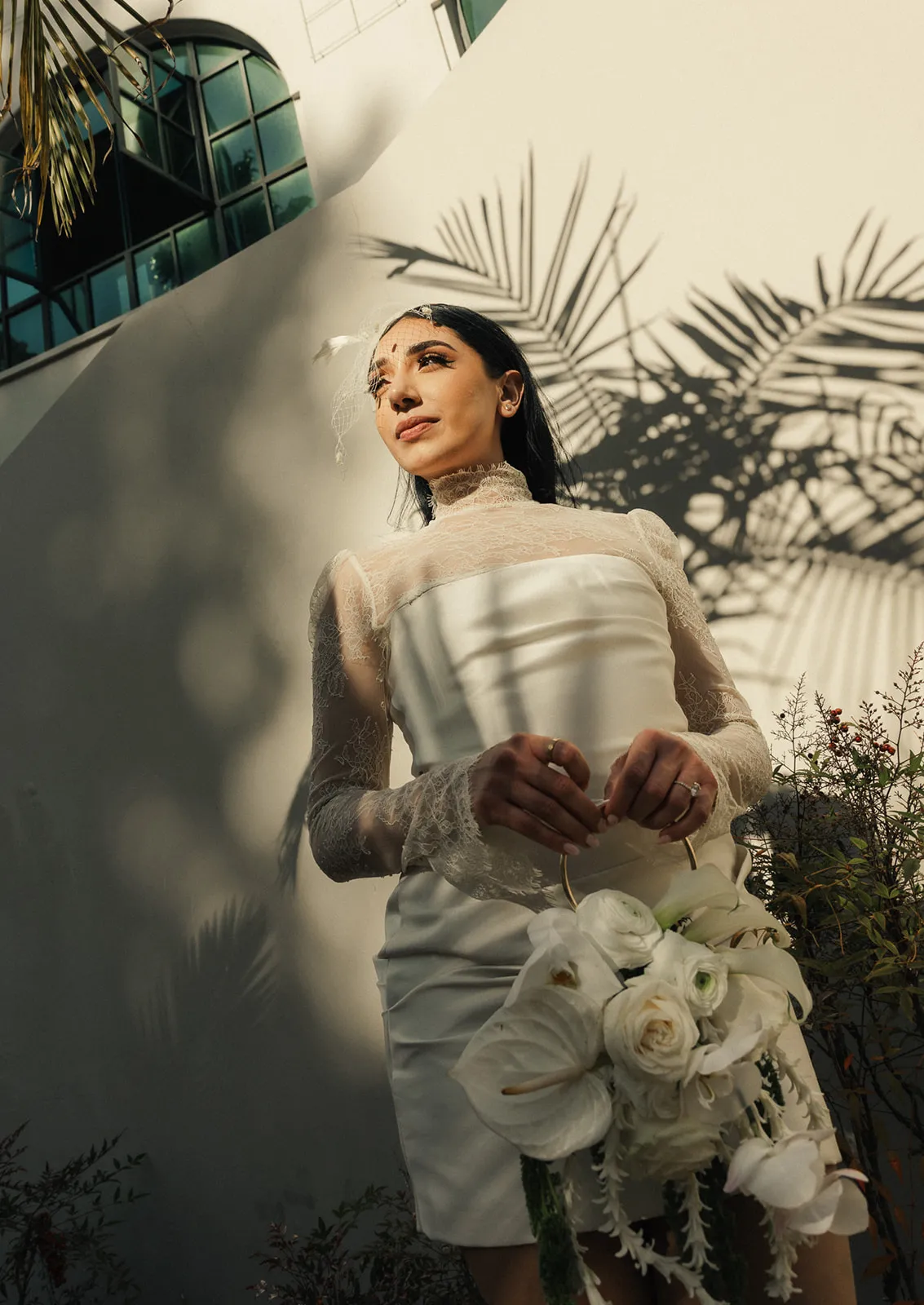 Low angle bride portrait against white wall with palm tree shadows at Beverly Hills City Hall, short lace turtleneck wedding dress and birdcage fascinator, holding white bouquet of roses and orchids, looking upward, photographed by Los Angeles elopement photographer Rene Porto