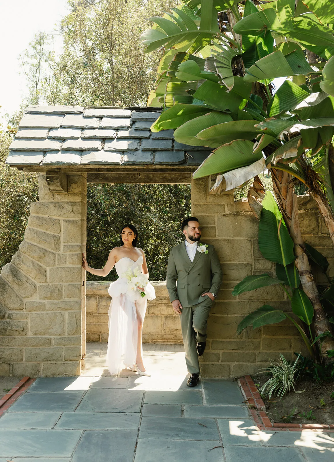 Couple portrait under a stone archway gate at Greystone Mansion Beverly Hills, bride in strapless white chiffon dress holding white orchid bouquet, groom in olive double-breasted suit leaning against the sandstone wall