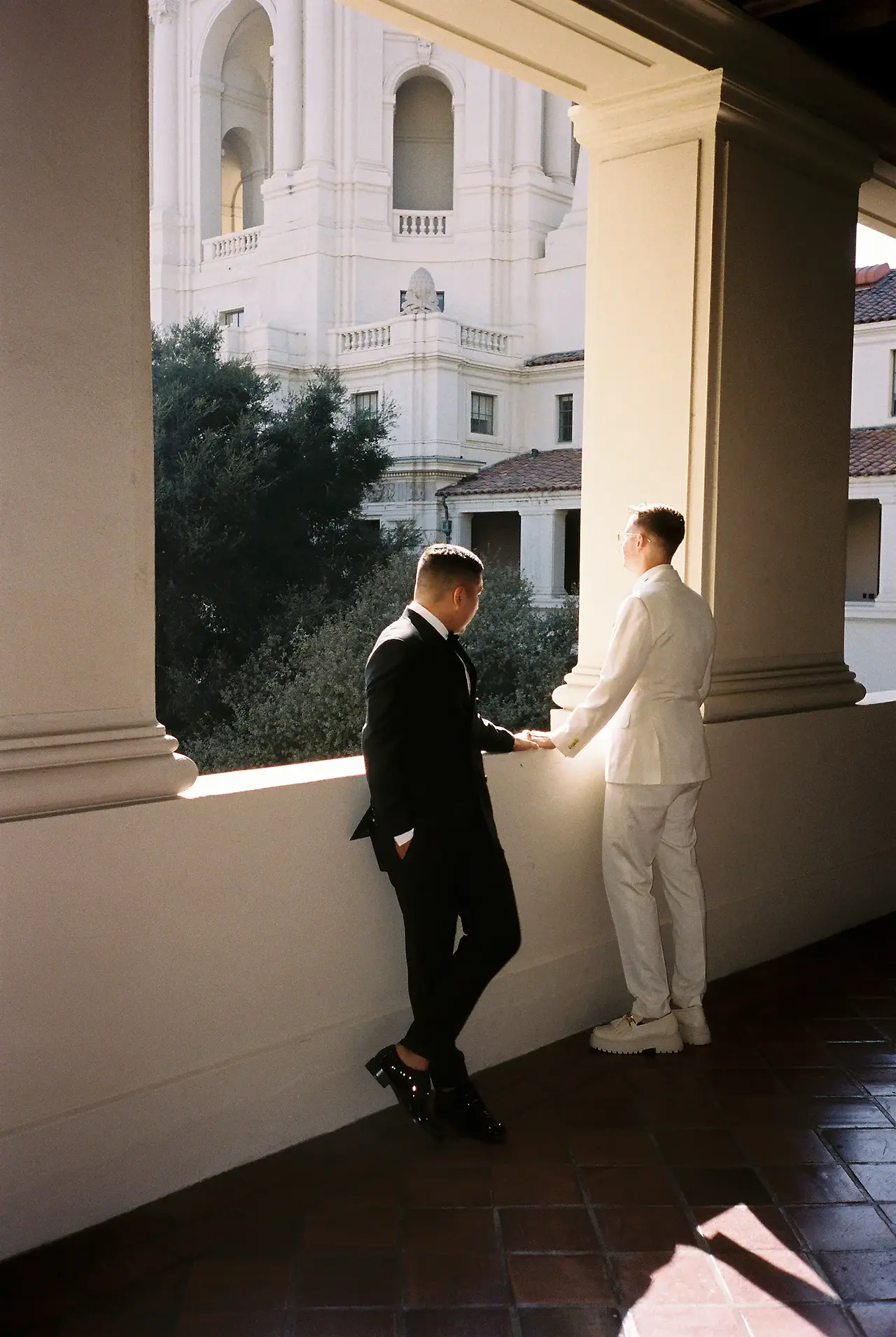 Two grooms walk along the arched loggia of Pasadena City Hall, one in a black tuxedo and one in a white suit, the dome and tower of the building visible through the columns, shot on 35mm film by Los Angeles film wedding photographer Rene Porto