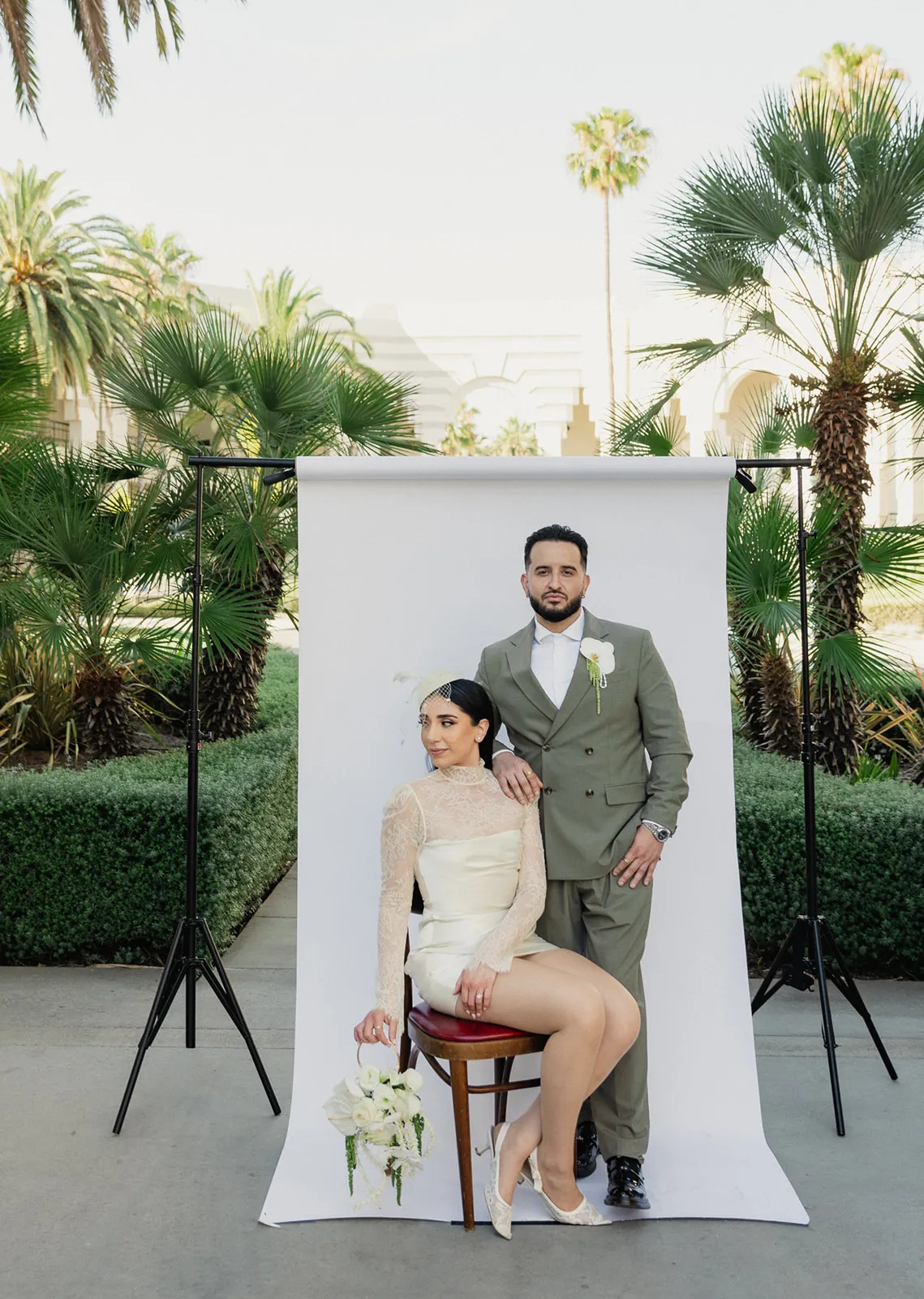 Couple posed against a white studio backdrop set up in the courtyard at Beverly Hills City Hall, bride seated on a bentwood stool in short lace turtleneck wedding dress and birdcage fascinator holding white calla lily bouquet, groom standing behind her in olive double-breasted suit with orchid boutonniere, City Hall arches and palm trees visible behind the setup, Beverly Hills courthouse wedding photography by Rene Porto