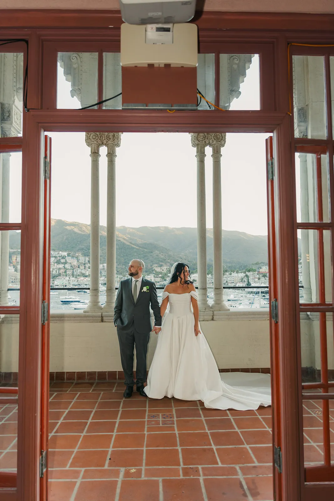 Bride and groom standing together framed by a red door and white ornate columns on the Casino veranda, full Avalon harbor view behind them