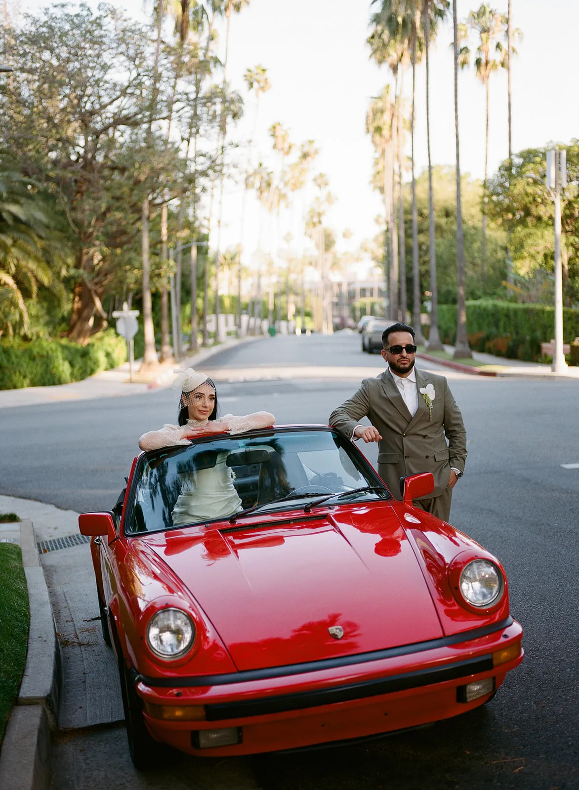 Couple with a vintage red Porsche 911 on a palm-lined Beverly Hills street at golden hour, bride seated inside the convertible in short lace dress and birdcage fascinator, groom leaning against the passenger door in olive double-breasted suit, shot on film by Los Angeles wedding photographer Rene Porto