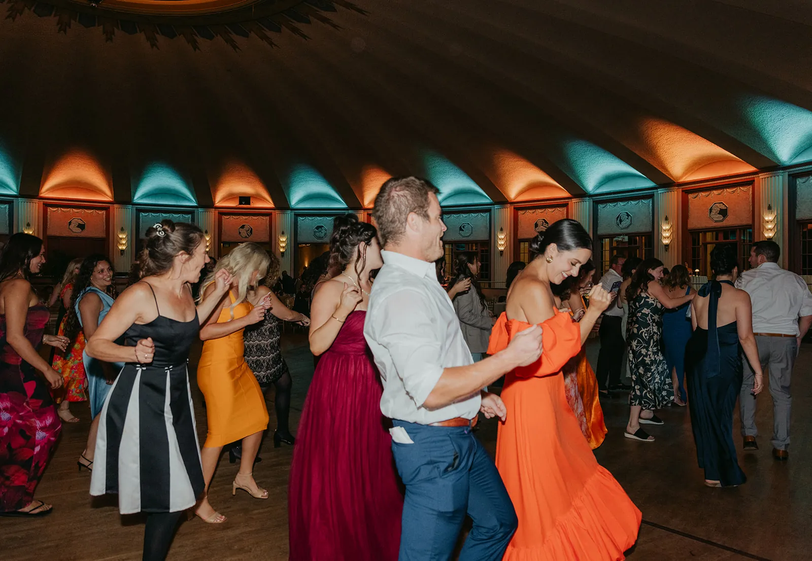 Full dance floor at the Catalina Island Casino Ballroom wedding reception with colorful gowns, live band on stage, and amber teal uplighting