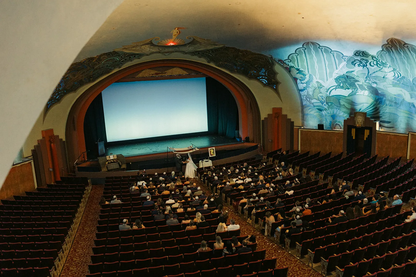 Aerial balcony shot of the full Avalon Theatre ceremony at the Catalina Island Casino, bride at the stage with the full crowd seated below and Beckman murals surrounding the room