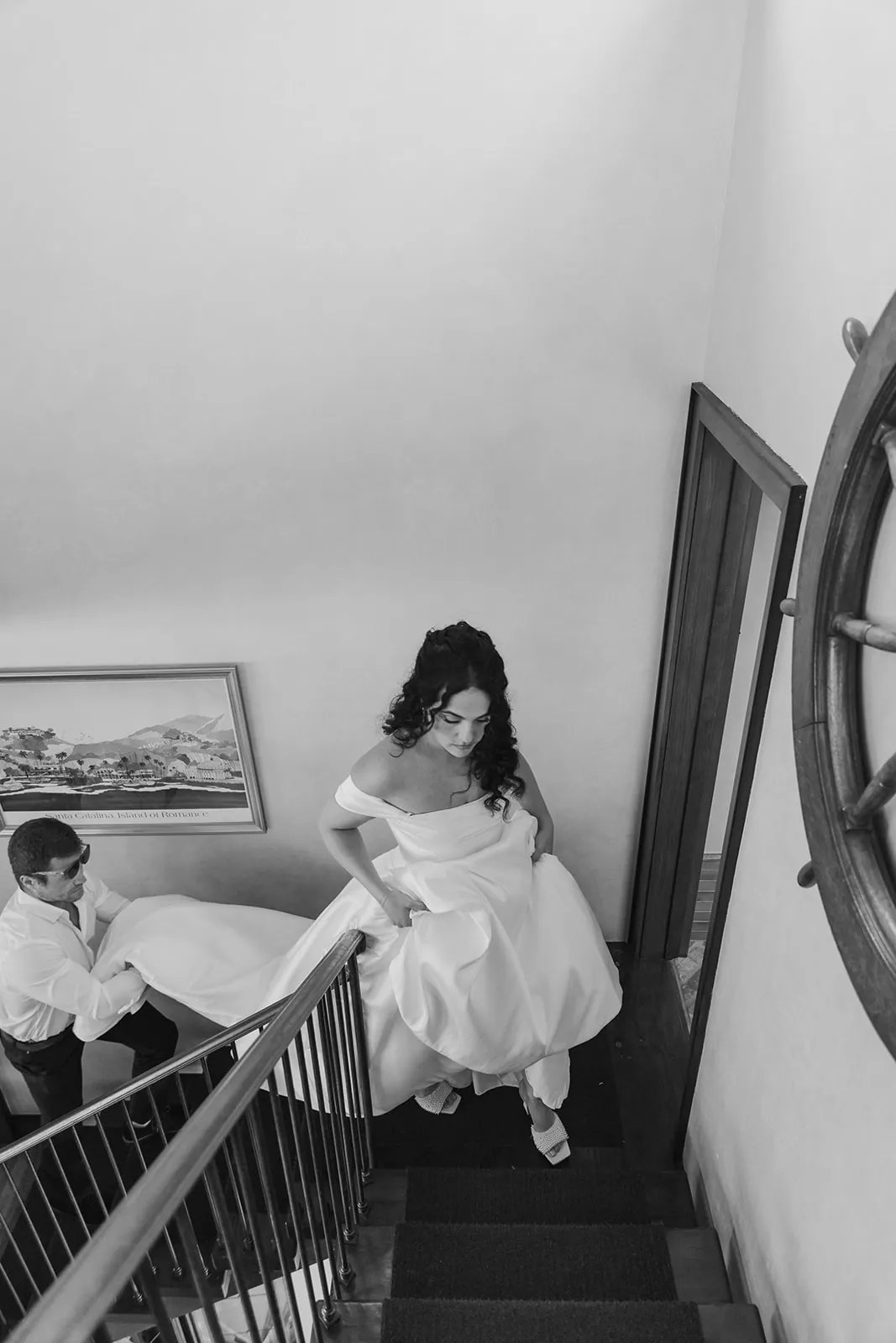 Bride pausing on the staircase beside a vintage Santa Catalina Island poster during getting ready at her Catalina Island Casino wedding, black and white