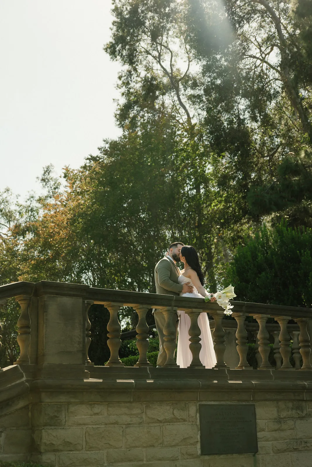 Couple kissing on the stone balustrade terrace at Greystone Mansion in Beverly Hills, bride in flowing white chiffon gown with cascading bouquet, groom in olive suit, photographed during a Beverly Hills courthouse wedding day by Rene Porto