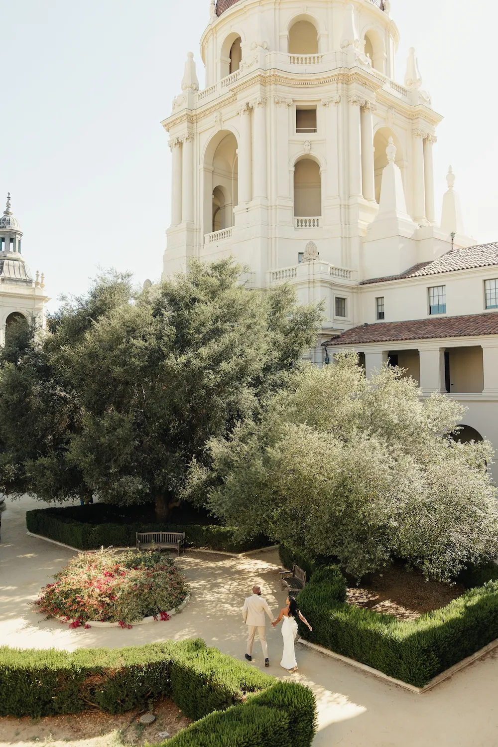 Aerial view of couple walking hand in hand through the Pasadena City Hall courtyard garden with the dome tower rising above them