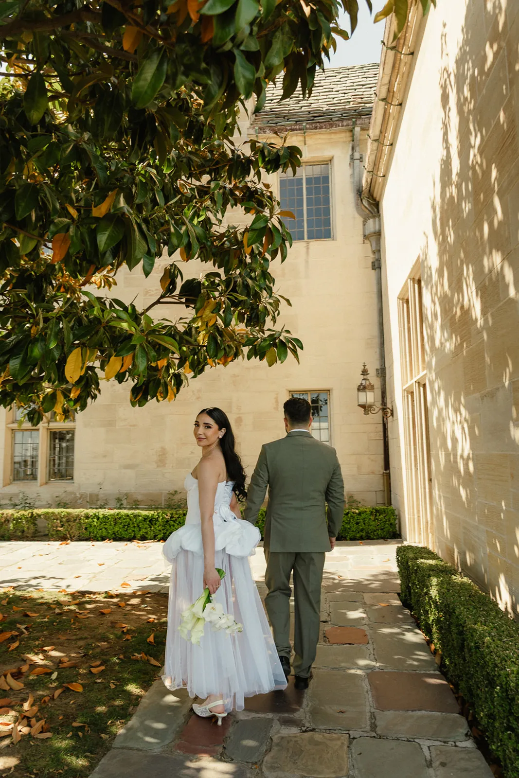 Couple walking along the sandstone wall of Greystone Mansion estate in Beverly Hills, bride in flowing white chiffon dress looking back over her shoulder with white bouquet, groom in olive suit leading the way, magnolia tree and dappled light overhead