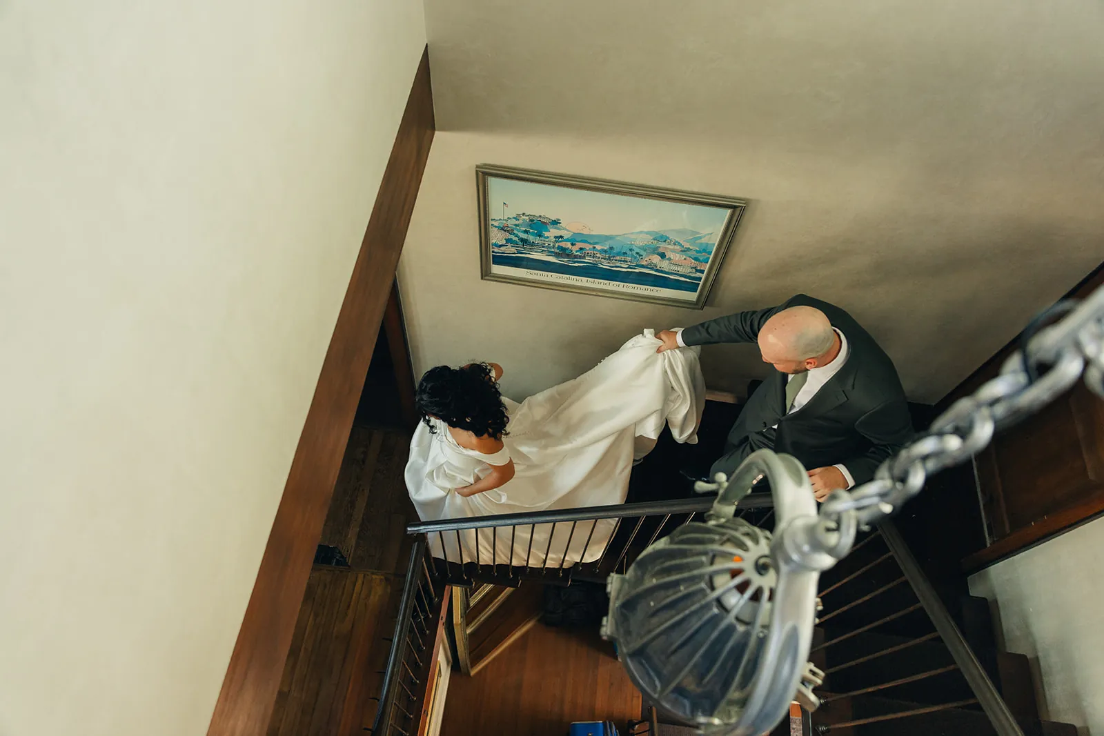 Overhead shot of bride descending the staircase with her train carried behind her, a vintage Santa Catalina Island painting visible on the wall, Catalina Island Casino wedding